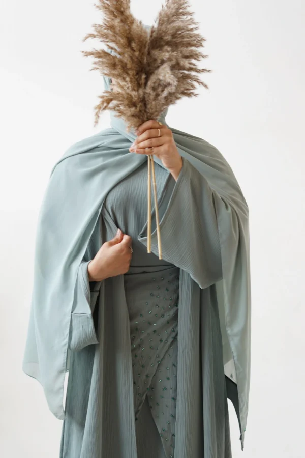 Person holding a bundle of pampas grass, partially obscuring their face, dressed in a textured sage green abaya with detailed beadwork.