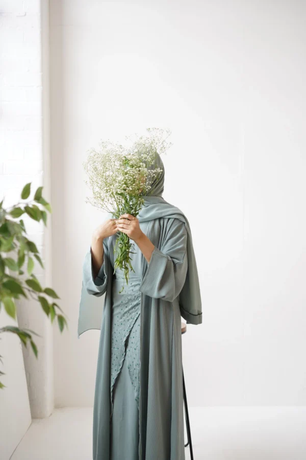 Person standing with a bouquet of baby's breath flowers covering their face, wearing a sage green abaya in a bright, airy room.