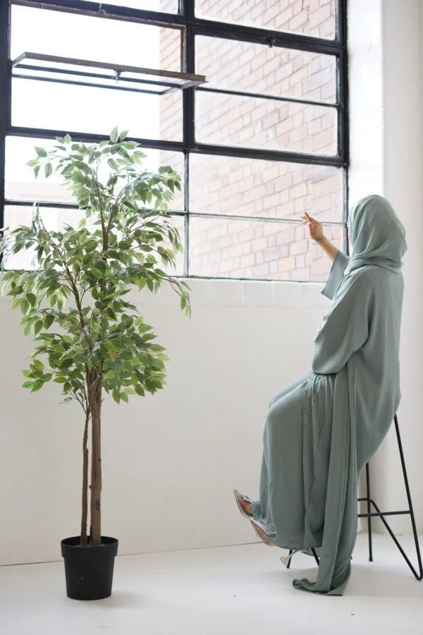Person in a sage green abaya looking out of a large window, creating a contemplative silhouette against the bright daylight.