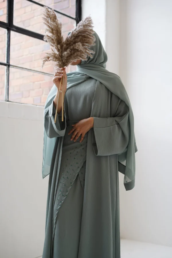 Person covered in a sage green abaya, holding pampas grass in front of their face, standing against a white wall with sunlight casting soft shadows.