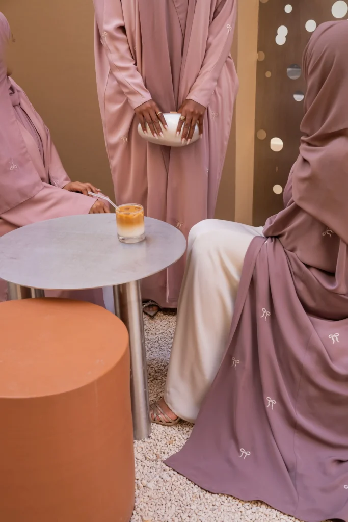 Three women in various styles of dusty rose bisht abayas with bow embroidery, seated and enjoying drinks at a modern café.